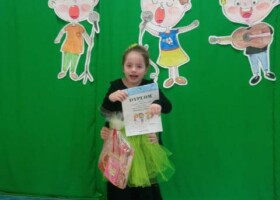 dziewczynka na tle zielonej dekoracji trzyma prezent i dyplom konkurs piosenki