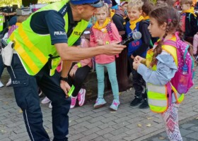 Na tle grupy dzieci policjant rozmawia z dziewczynką do mikrofonu